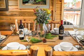 Una mesa de madera con un florero de flores, dos botellas de vino y vajilla blanca.