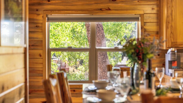 A wooden interior with a dining table set for four, a window with a view of greenery, and a kitchen area with appliances.