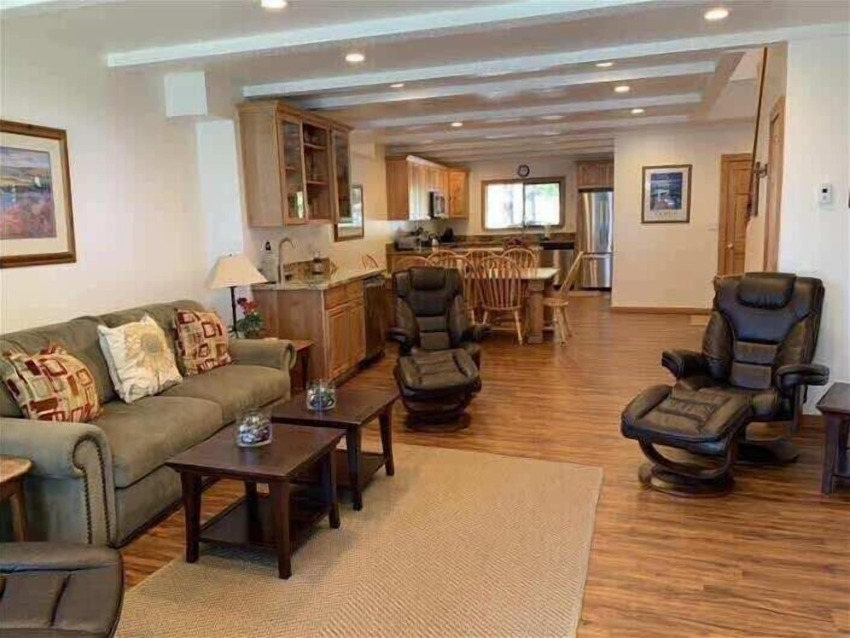 A spacious living room with a sofa, armchairs, and a coffee table. There is a kitchen area with wooden cabinets and a dining table.