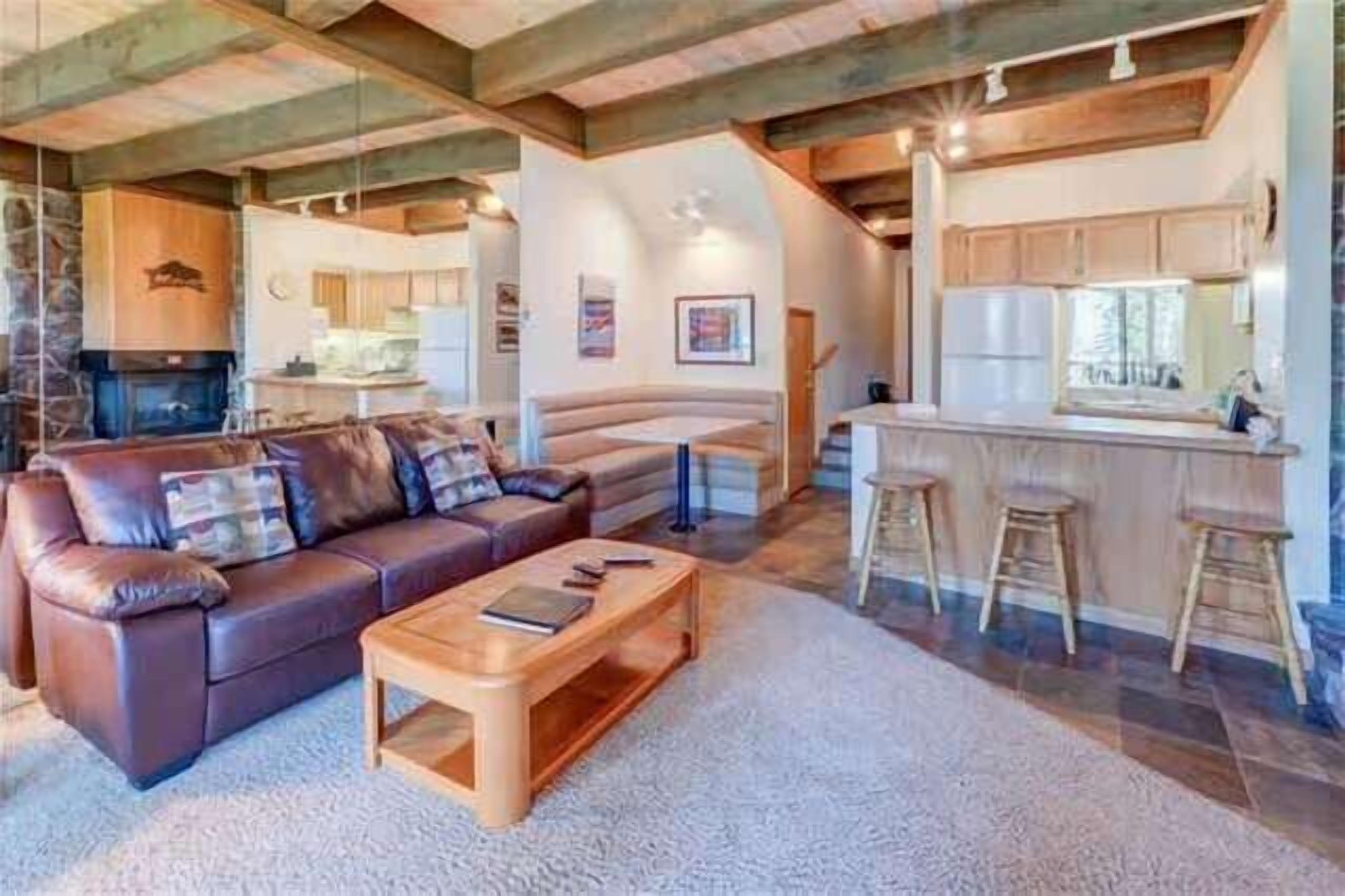 A cozy living room with a leather sofa, wooden coffee table, and a fireplace. There is a kitchen area with wooden cabinets and a bar stool.