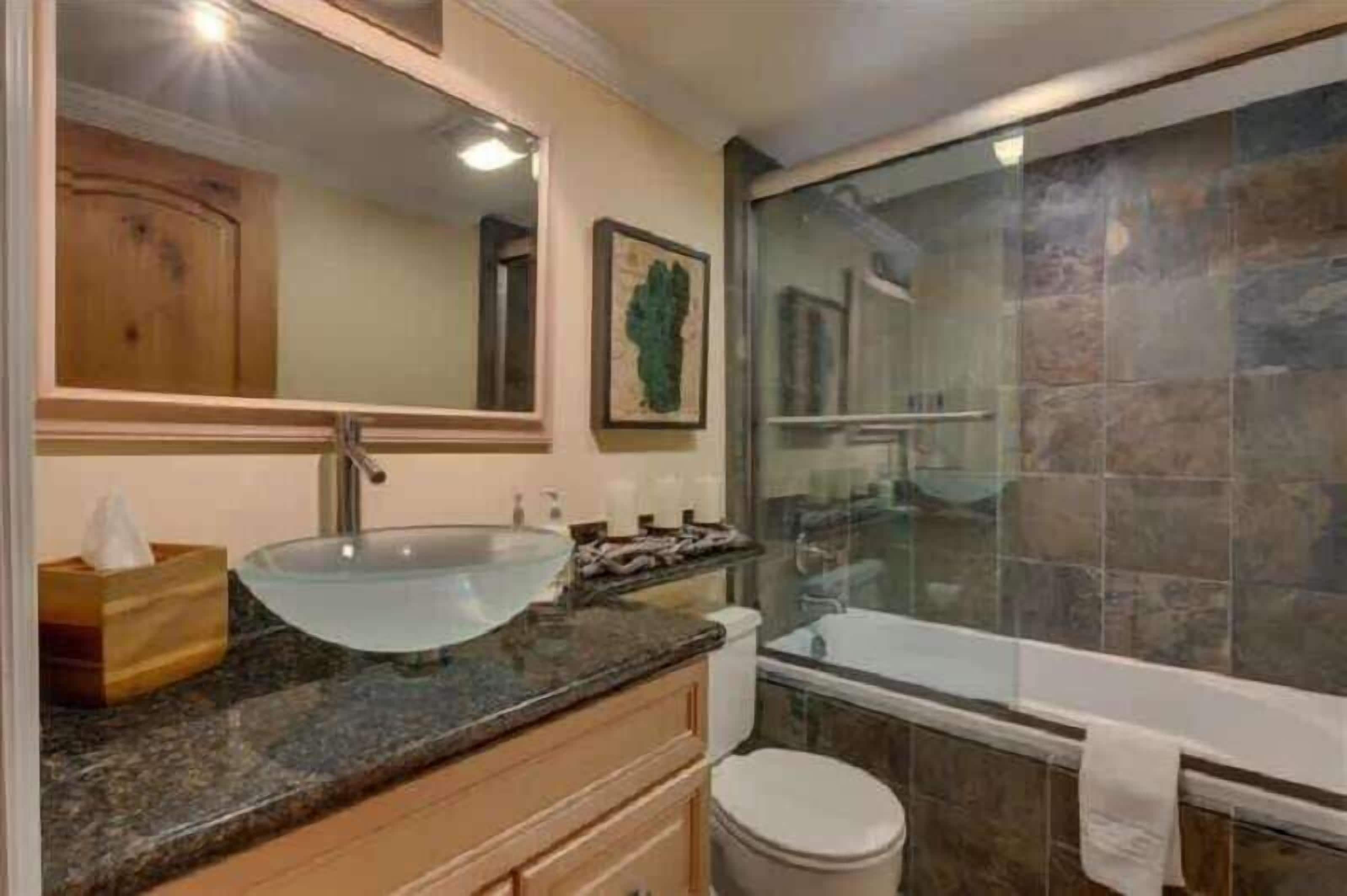 A bathroom with a glass sink, wooden vanity, a large mirror, and a bathtub with a stone wall.