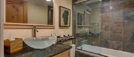 A bathroom with a glass sink, wooden vanity, a large mirror, and a bathtub with a stone wall.