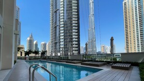 A rooftop pool with a metal railing, surrounded by a wooden deck and lounge chairs, with high-rise buildings in the background.