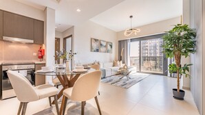 A modern kitchen with a built-in oven, a dining area with a glass table, and a living room with a sofa and a large window.