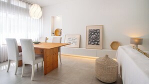 Et moderne, minimalistisk værelse med et træspisebord, hvide stole, en vævet puf, en seng med hvide sengetøj og kunst på væggene.