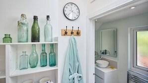 Un baño con un reloj, estantes con botellas de vidrio, un perchero para toallas y un lavabo con espejo.