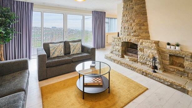 Una sala de estar con chimenea de piedra, un sofá gris, una mesa de centro de vidrio y vistas amplias a través de grandes ventanales.