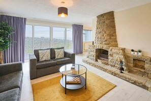 Una sala de estar con chimenea de piedra, un sofá gris, una mesa de centro de vidrio y vistas amplias a través de grandes ventanales.
