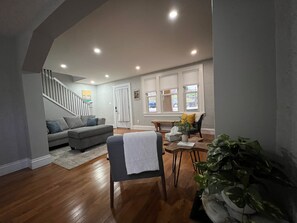 A spacious living room with a grey sofa, a wooden coffee table, and a dining table with chairs. A staircase leads to an upper level.