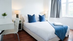 Een moderne slaapkamer met een wit bed, blauwe kussens, een bureau en een stoel.