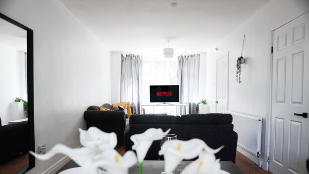Een moderne woonkamer met een bank, een eettafel met witte borden en bloemen, een tv waarop 'NETFLIX' wordt weergegeven, en een witte deur.