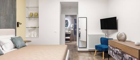 A modern bedroom with a bed, a dresser, a TV, and a bathroom visible through an open door.
