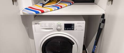 A compact laundry area with a front-loading washing machine, a shelf with an iron and a safe, and a mop leaning against the wall.