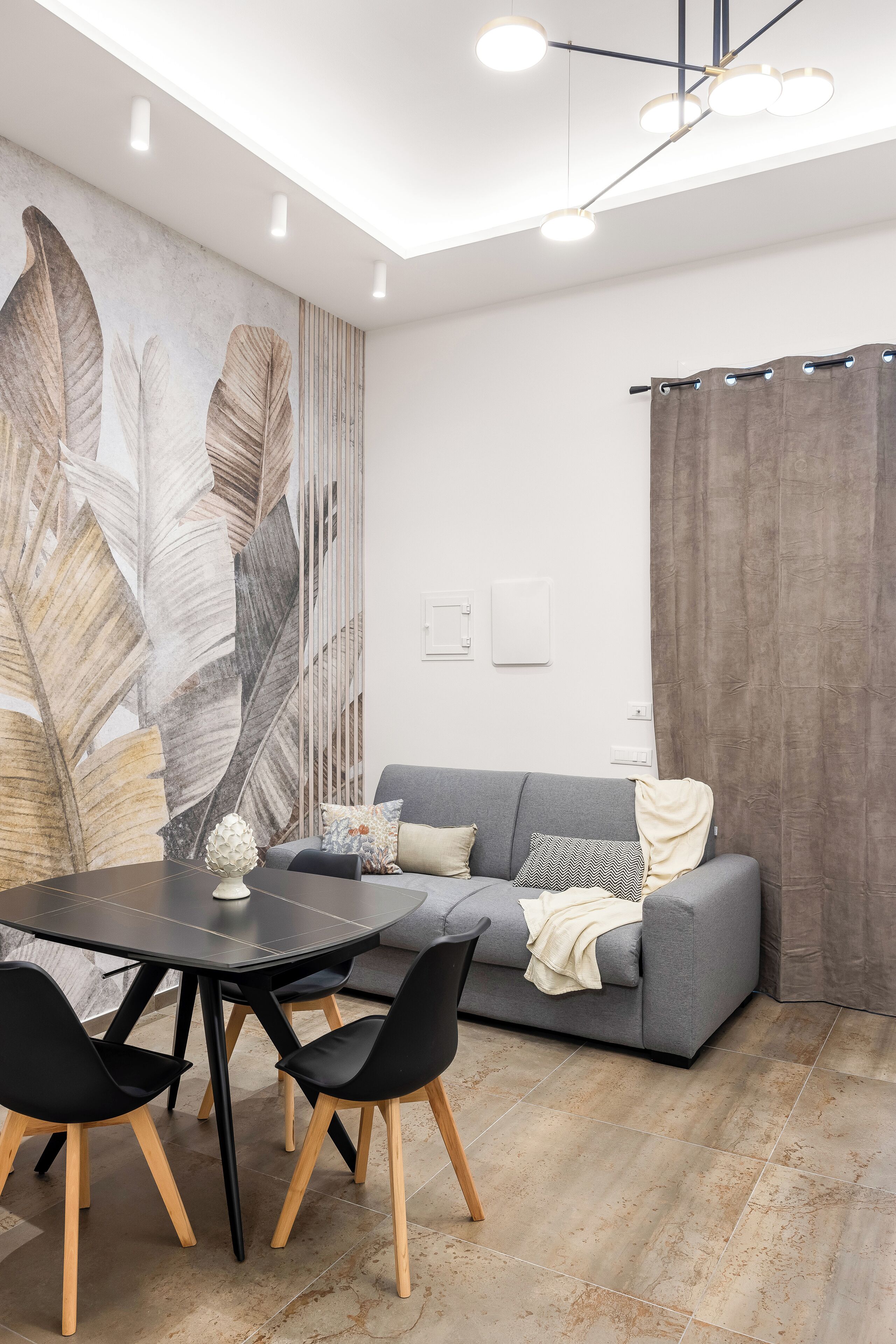 A modern living room with a grey sofa, a dining table with black chairs, and a large leaf-patterned wall mural.