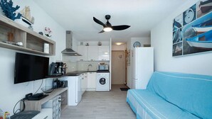 A compact living space with a kitchenette, a washing machine, a flat-screen TV, a blue sofa, and a ceiling fan. - Apartment in Agde near Beach & Heated Pool (Agde)