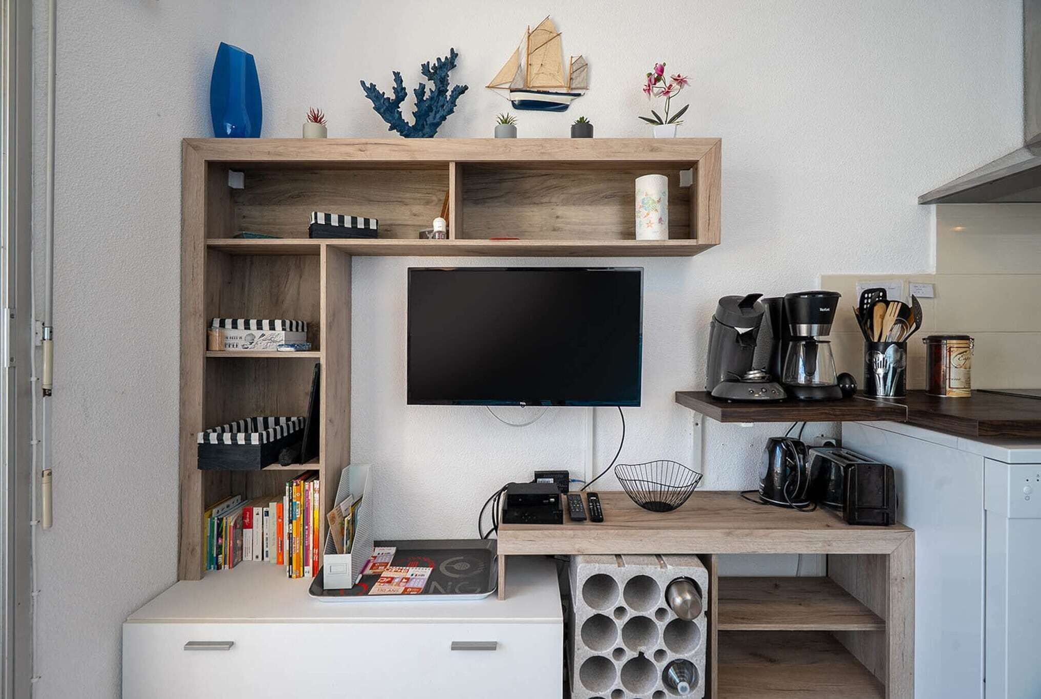 Un espace de vie compact avec un téléviseur fixé au mur, un bureau en bois avec une machine à café et une bibliothèque remplie de livres.