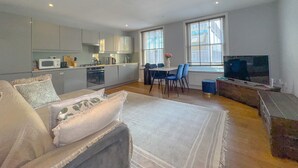 A modern living room with a sofa, a coffee table, and a TV. There is a dining area with a table and chairs. The kitchen is visible with appliances and cabinets. - Pass the Keys | Central 2 Bed 2 Bath Duplex (London)