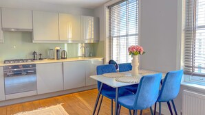A modern kitchen with white cabinets, a dining area with blue chairs, and a large window with blinds. - Pass the Keys | Central 2 Bed 2 Bath Duplex (London)