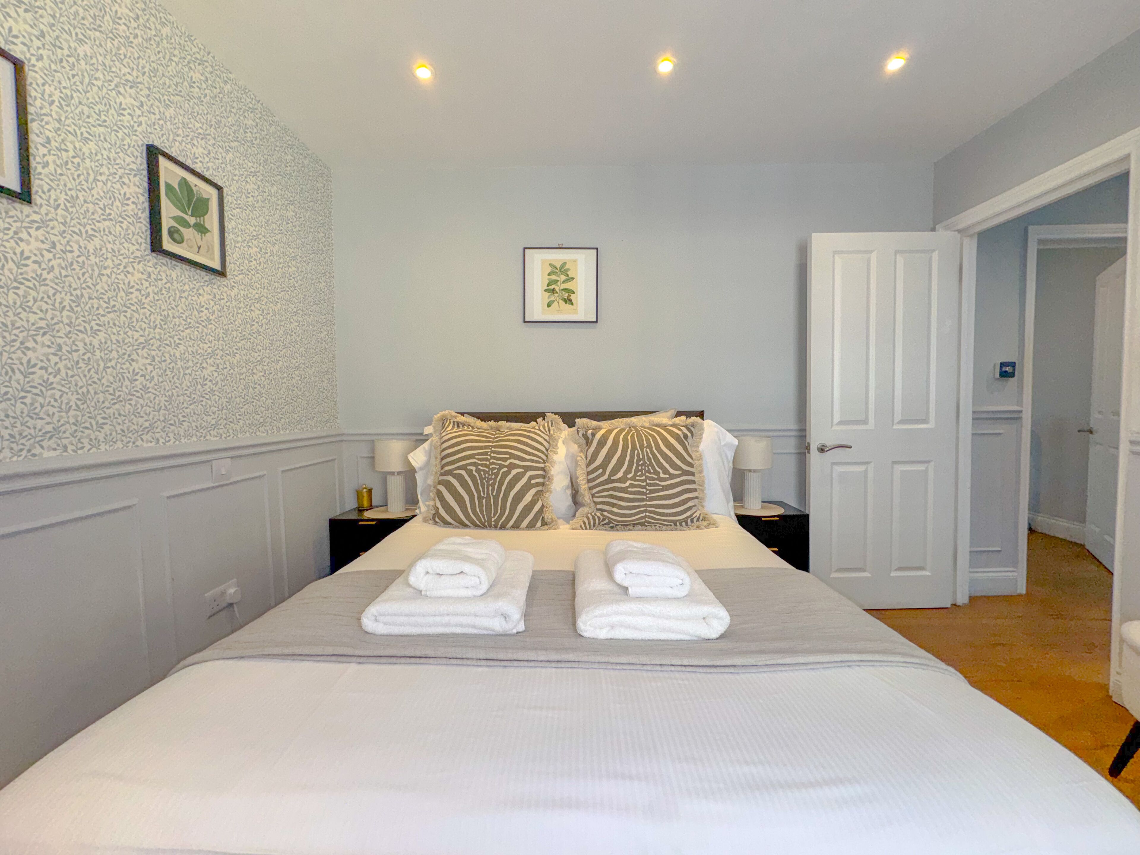 A neatly made bed with two folded towels, a zebra-patterned pillow, and a framed botanical print on the wall.