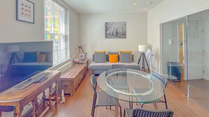 Habitació moderna amb taula de dinar de vidre, un sofà amb coixins, un llit i una finestra amb persianes.