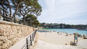 A stone staircase leading to a beach with a railing, a clear blue sea, and buildings in the background. - Banc Doli - Villa With Private Pool In Manacor (Manacor, Illes Balears)