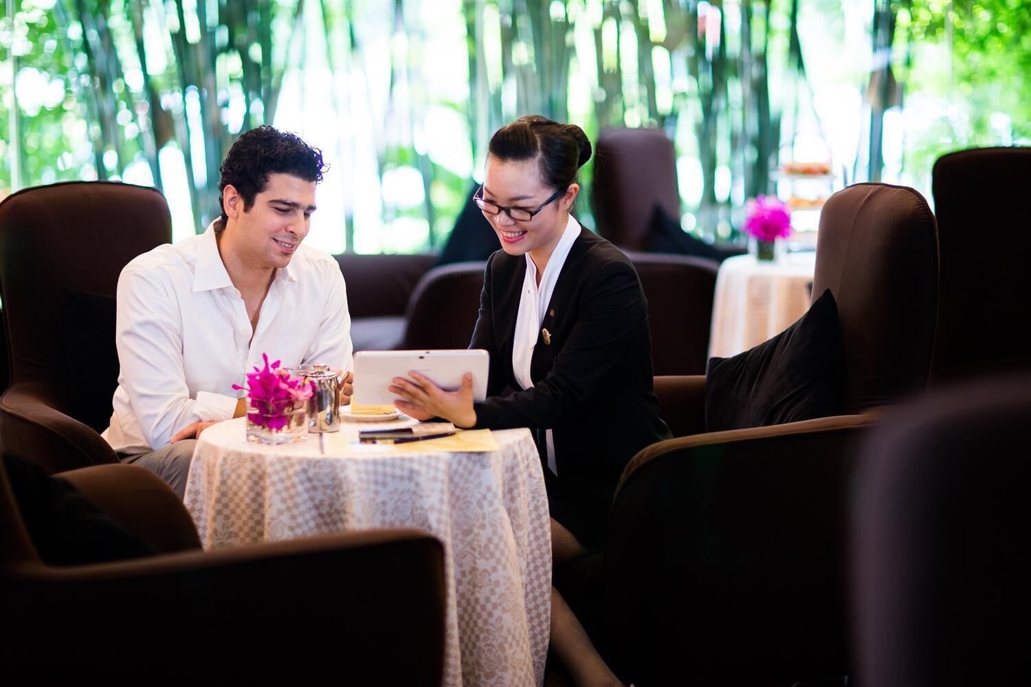 Two people seated at a table with a laptop, in a room with a view of greenery through large windows.