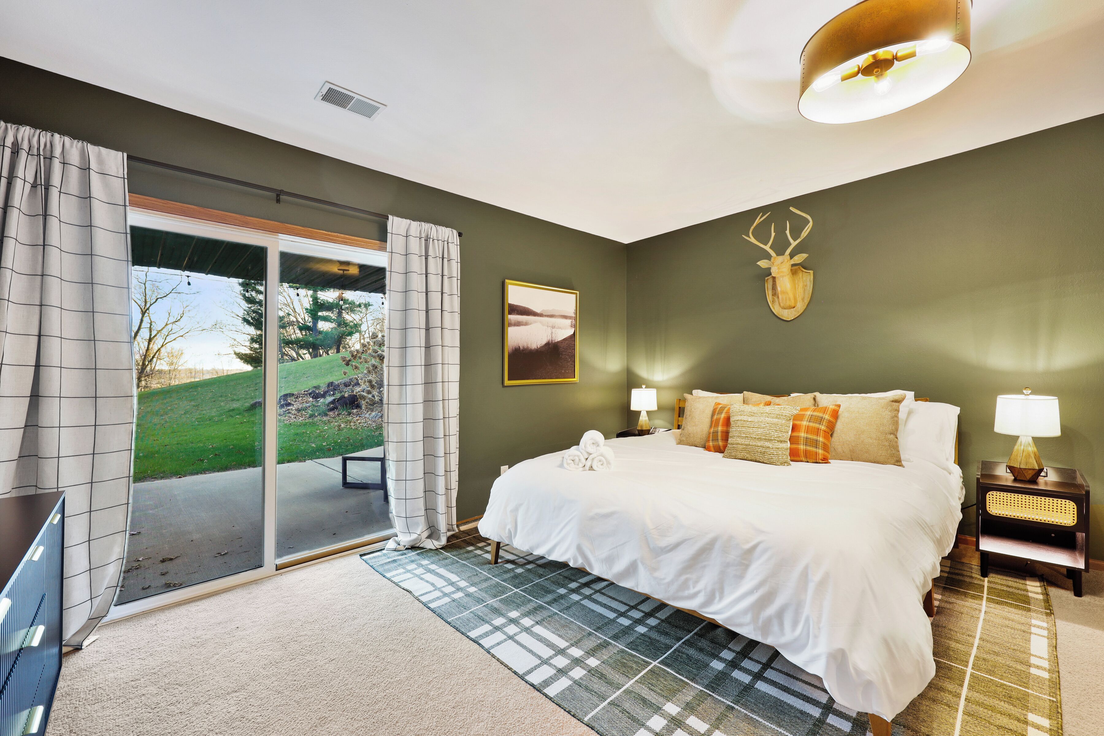 Une chambre avec un grand lit, une vue à travers une porte-fenêtre coulissante, une tête de cerf accrochée au mur et une lampe sur une table de chevet.