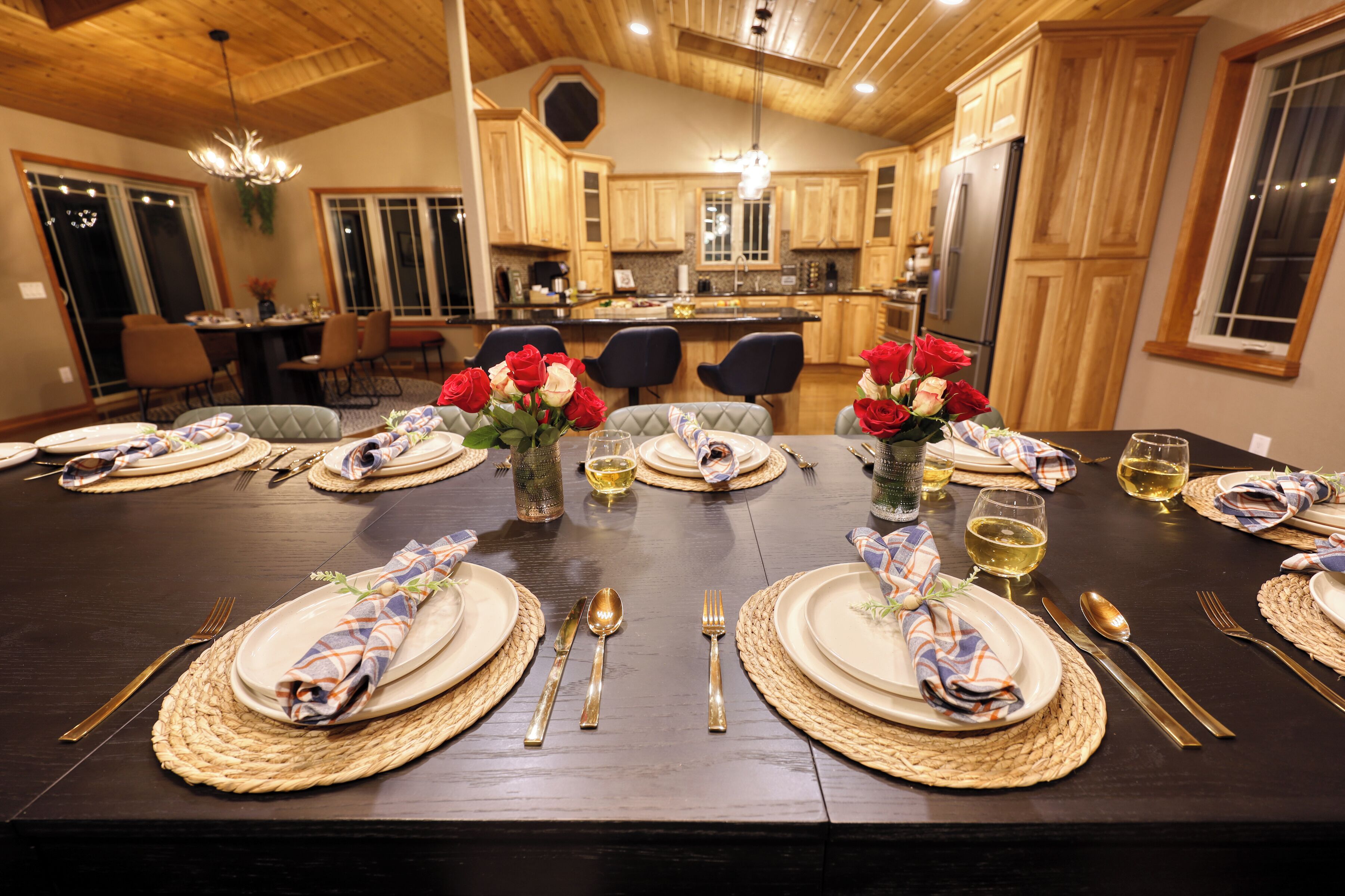 A dining table set with plates, cutlery, and wine glasses, surrounded by wooden cabinets and a chandelier.