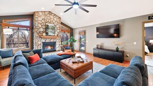 A spacious living room with a stone fireplace, large windows, and a ceiling fan. - Wild Timber Lodge: Golf Sim, Sauna, Bars, 10 acres, close to WI Dells (Oxford)