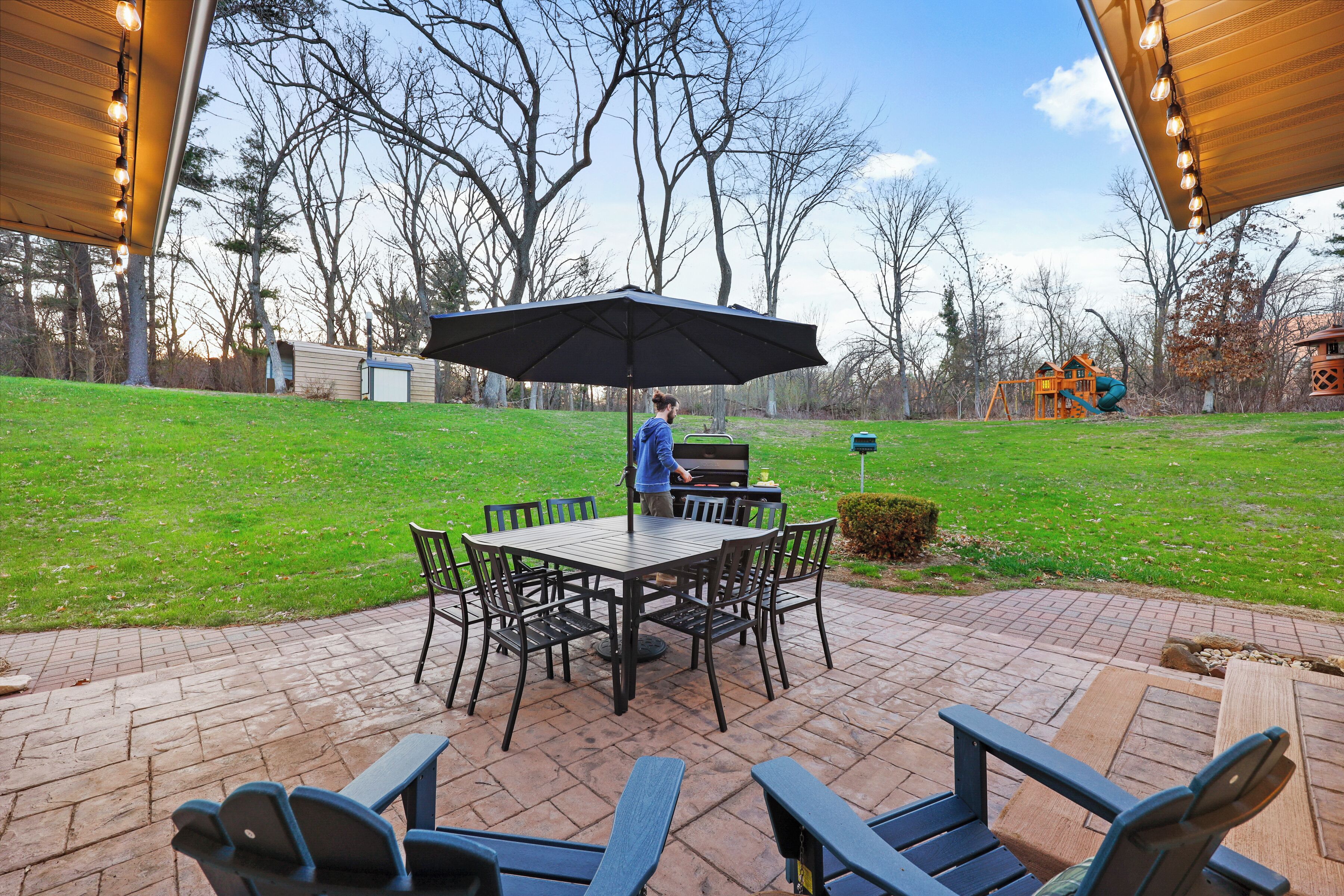 Une terrasse extérieure avec une table et des chaises, un barbecue et, au fond, un ensemble de jeux pour enfants avec une balançoire.