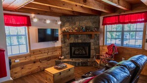 A cozy cabin interior with a stone fireplace, wooden walls, a leather couch, and a television.