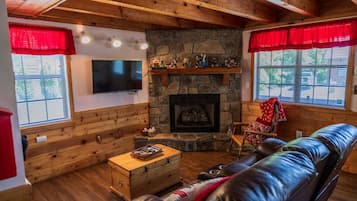 A cozy cabin interior with a stone fireplace, wooden walls, a leather couch, and a television.