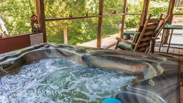 A hot tub with a wooden deck and chairs, surrounded by trees.