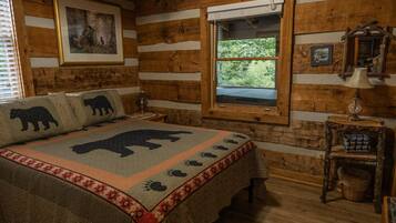 A log cabin bedroom with a bed featuring a bear-themed blanket, a window with a view of greenery, and a rustic wooden wall.