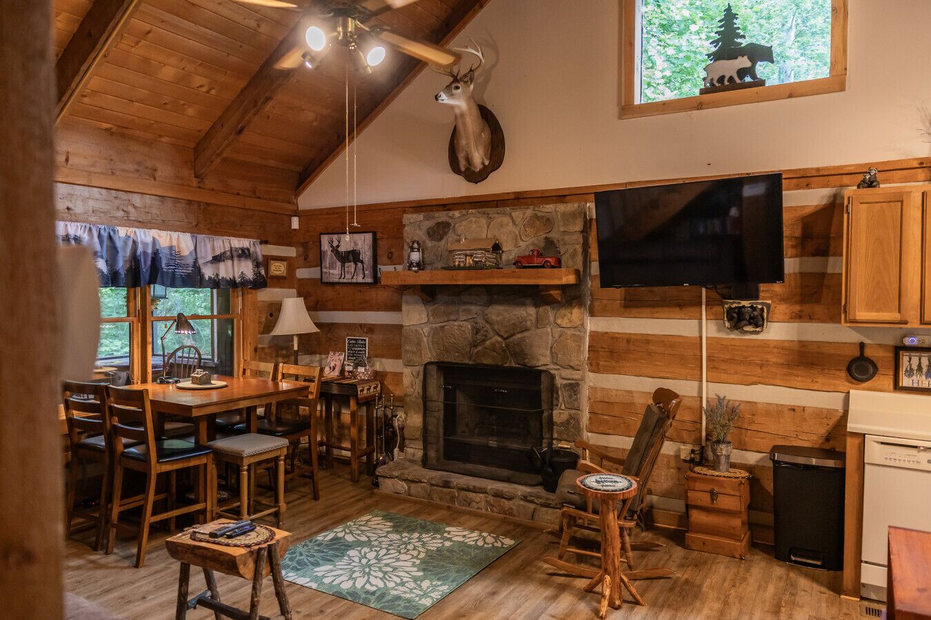 A cozy cabin interior with a stone fireplace, wooden furniture, a mounted deer head, and a television.
