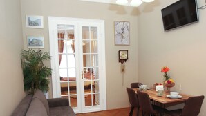 A living room with a sofa, a dining table with chairs, a TV mounted on the wall, and a door leading to another room.