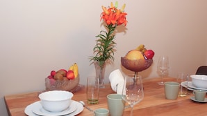 A wooden dining table with a bowl of fruit, a vase with flowers, and various cups and glasses.