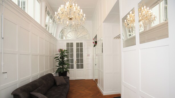 A hallway with a dark brown sofa, a potted plant, and a large mirror reflecting a chandelier.