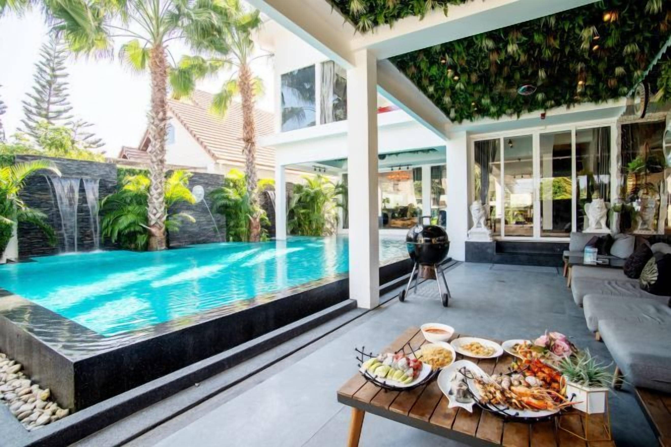 A poolside area with a dining table set with seafood, a grill, and a view of a waterfall and palm trees.