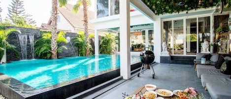 A poolside area with a dining table set with seafood, a grill, and a view of a waterfall and palm trees.