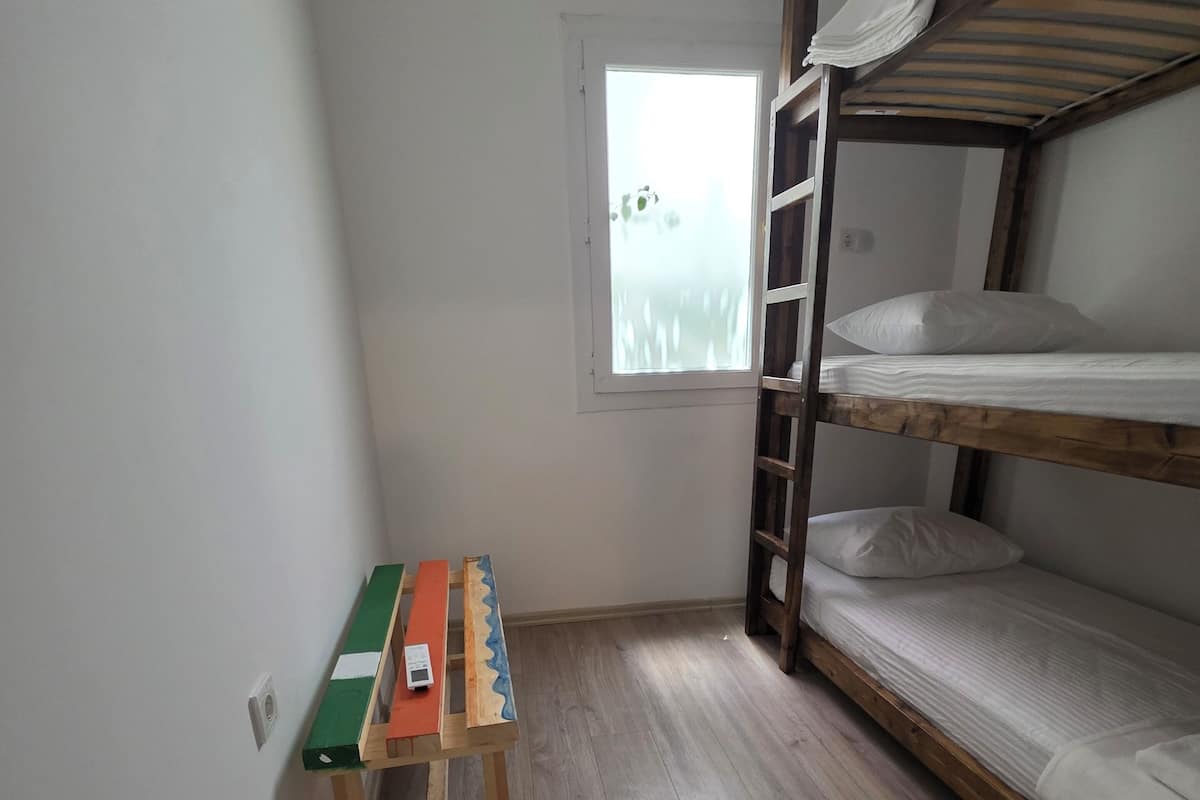 Eine kleine, einfache Unterkunft mit Etagenbett, einem Holztisch mit Stuhl, einem Fenster mit Milchglas und einer Klimaanlage an der Wand.