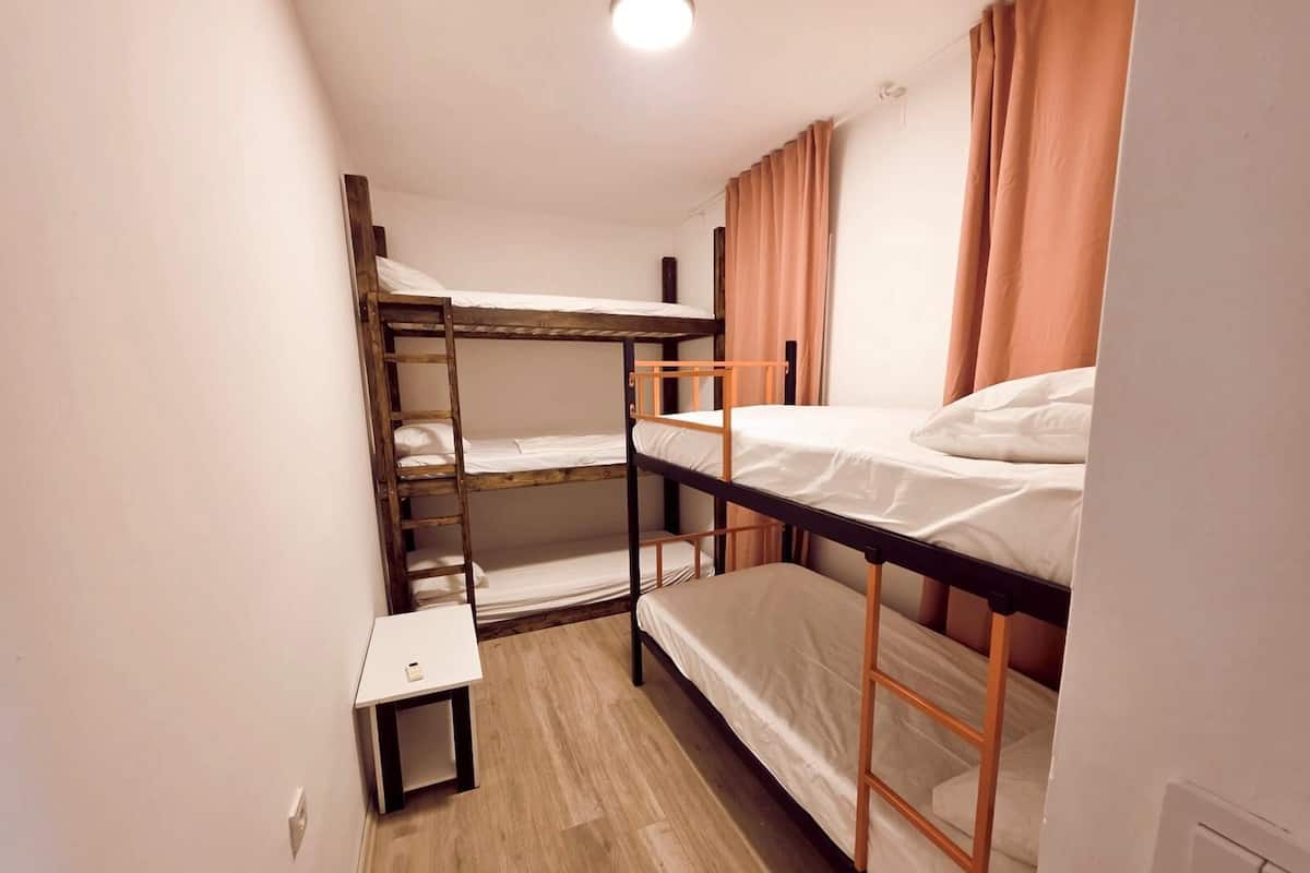 Ein kleines, schlichtes Zimmer mit Etagenbetten, einem kleinen Tisch und einer einzelnen Lampe.