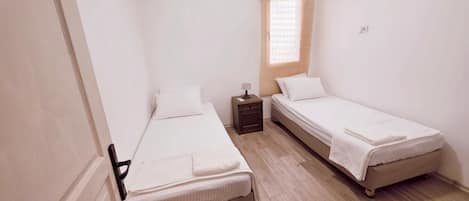 A small, clean room with two single beds, a nightstand, a window with blinds, and an air conditioning unit.