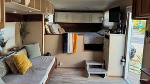 Un espai habitable reduït amb sofà, taula petita, zona de cuina i finestra amb persianes.