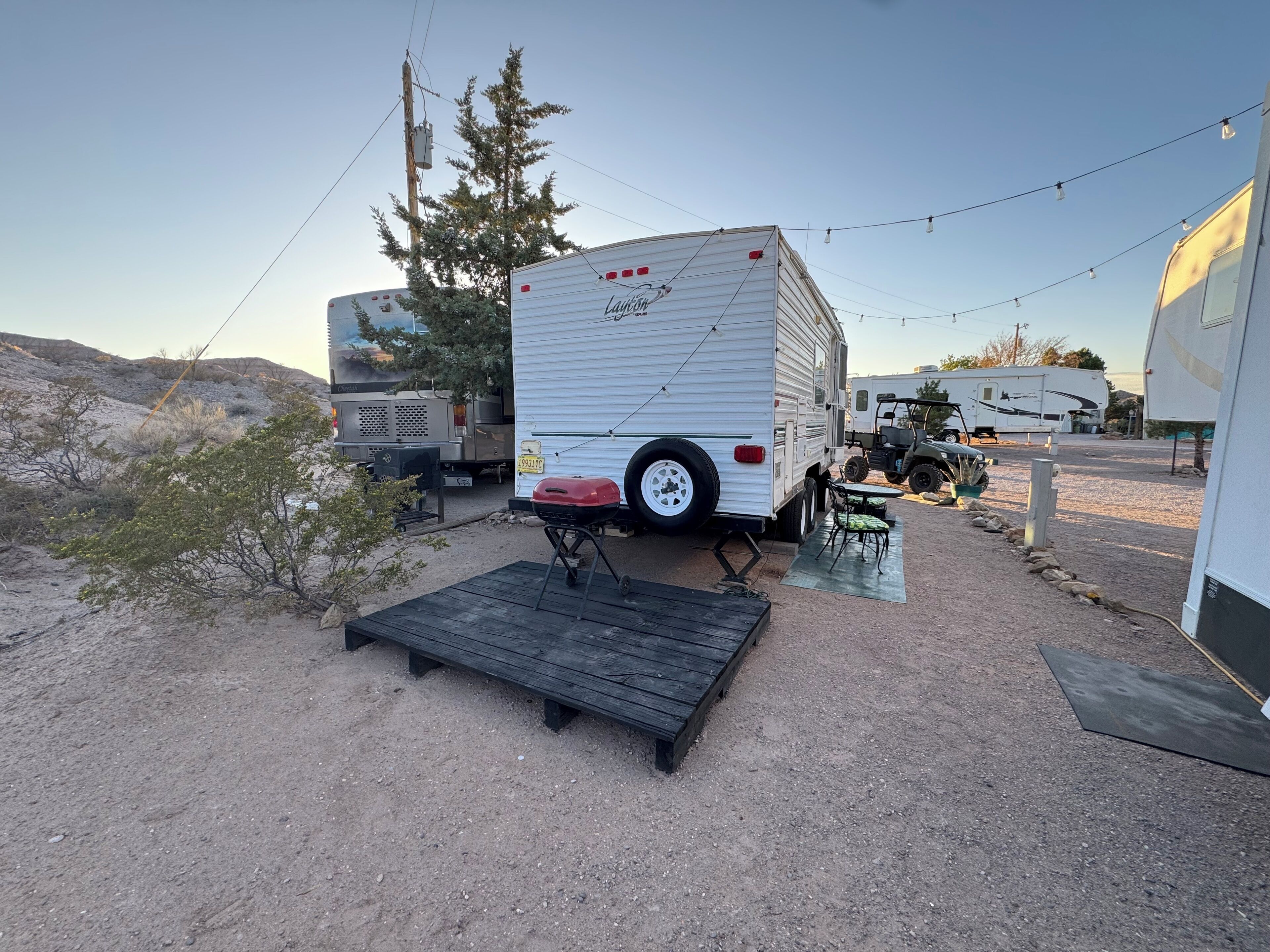 A campsite with a white RV named "Layton" parked on a gravel surface, a wooden deck, and a small table with chairs.