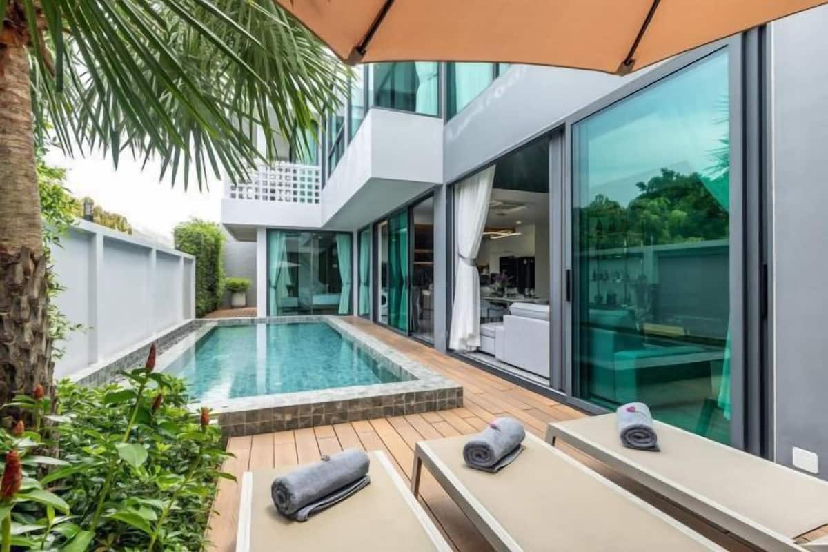 Un área de piscina moderna al aire libre con un porche de cristal, sillas de descanso y una palmera.