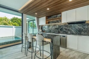 A modern kitchen with a marble countertop, white cabinets, and a built-in island with bar stools. - Mandla Villas Collection-Villa Tanya (Phuket)
