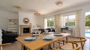 Un comedor con una mesa de madera dispuesta para cuatro personas, rodeada de sillas de mimbre. Hay una chimenea con repisa, una estantería y vistas a una piscina a través de amplias ventanas.