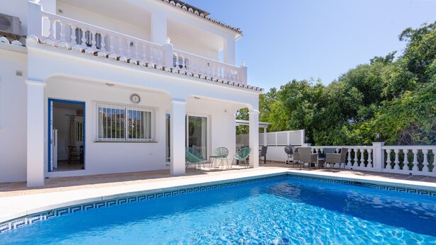 Una casa de dos pisos con piscina, mobiliario exterior y cerca blanca.