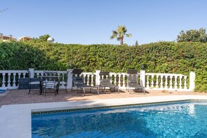 A swimming pool with lounge chairs and a table, surrounded by a white fence and lush greenery. - Villa Blue Skies - Calahonda (Sitio de Calahonda)